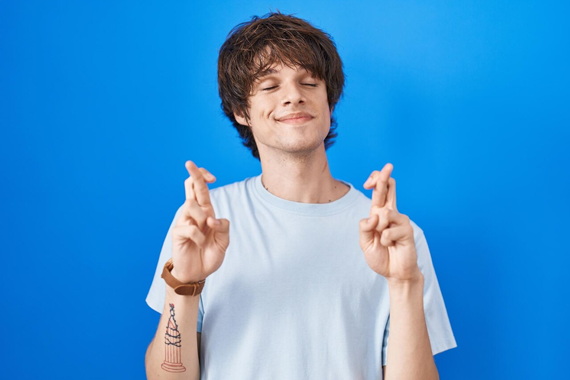 Kurz vor dem TMS Männliche weiße Person mit dunklen mittellangen Haaren und weißen T-Shirt hat die Augen geschlossen und überkreuzt Mittel- und Zeigefinger beider Hände. Der Hintergrund ist eine blaue Wand.